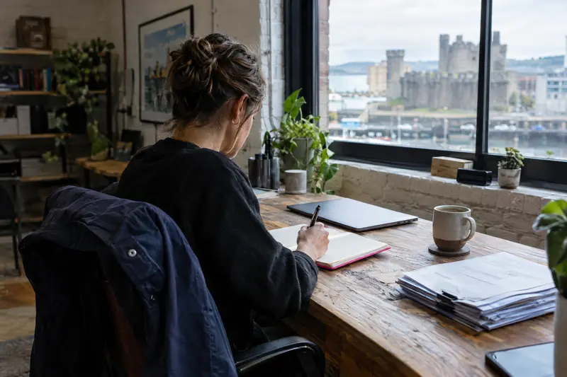 Small business owner at a workshop desk writing a task list beside a closed laptop and coffee