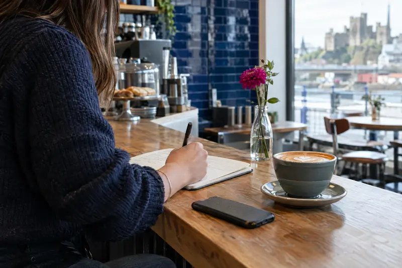 Café owner writing article notes in a notebook while a phone lies face down on the counter