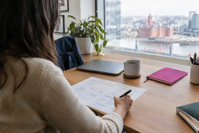 Small business owner at a wooden desk reviewing a printed website wireframe with a pen beside a laptop